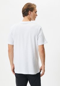 Valkoinen puuvillapaita lyhyillä hihoilla, pyöreällä kaulahalkaisijalla ja sileällä pinnalla, esiteltynä takaviistosta. Ei näkyviä logoja tai kuvioita.