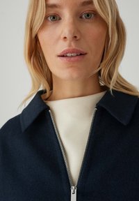 Giacca in misto lana blu navy con colletto appuntito e chiusura lampo argento, caratterizzata da una superficie testurizzata ed elementi di design minimalisti.