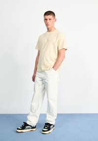 Beige katoenen t-shirt met reliëflogo, gecombineerd met witte cargo-broek met zakken en gouden hardware. Zwart-witte sneakers maken de look compleet.