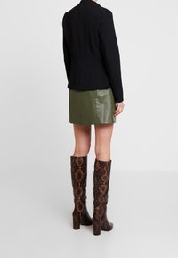 Czarna, teksturowana marynarka, zielona mini spódnica ze skóry oraz brązowe kozaki za kolano z wzorem wężowej skóry i grubym obcasem. Prosty, przemyślany design.