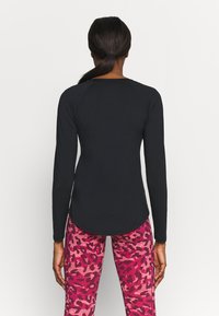 Woman with dark hair in a ponytail wears a fitted long-sleeve black top and pink camo-patterned leggings, standing facing away against a plain background.