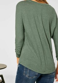 Grön långärmad topp med ribbad textur, med en avslappnad passform, rundad halsringning och rynkade detaljer på ärmarna, tillsammans med blå jeans.