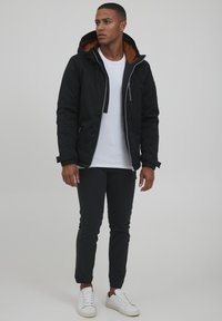 Black insulated jacket with a brown interior lining, zip closure, and hood. Paired with black pants and white sneakers.