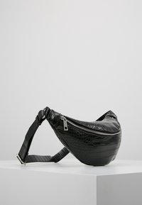 Svart fanny pack av läder med krokodilsmönster, med dragkedjestängning, justerbar rem och en böjd form, vilande på en vit yta.