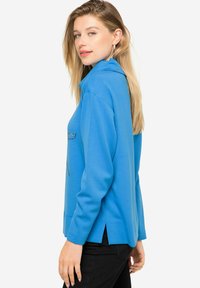 Sudadera de color azul claro con cuello, aberturas laterales y bolsillo frontal. Fabricada con tela suave, tiene mangas largas y un corte relajado.