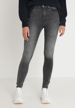 Persoon die zwarte gewassen skinny jeans, witte sneakers en een donker geribde trui draagt, staand tegen een effen achtergrond.