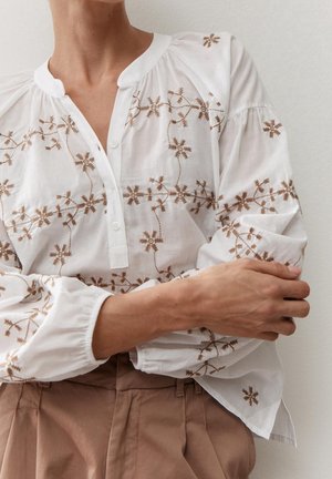 Woman wearing a white blouse with brown floral embroidery and beige pants, arms crossed, against a plain light background.