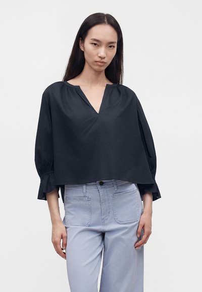 Jeune femme aux longs cheveux foncés portant un chemisier noir ample à manches trois-quarts et un pantalon taille haute bleu clair sur un fond uni.