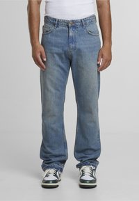 Muž nosiaci voľné, vyblednuté modré rifle, biele tielko a bielo-čierno-zelené tenisky stojaci pred jednoduchým pozadím.