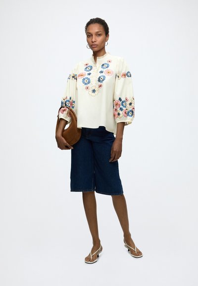 Femme portant un chemisier crème avec une broderie florale colorée et un short en denim foncé longueur genou, tenant un sac à main marron et portant des sandales blanches.