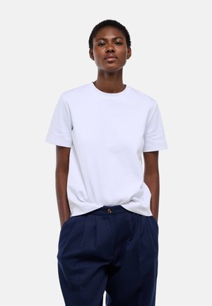 Femme aux cheveux courts portant un t-shirt blanc uni à manches courtes et un pantalon ample bleu foncé, debout les mains dans les poches.