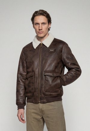 Homme portant une veste en cuir marron avec col en peau lainée blanche, fermeture éclair devant, poches à rabat et pantalon beige, debout sur un fond uni.