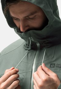 Giacca impermeabile verde con colletto alto, cordino regolabile e logo del marchio. Texture liscia e chiusura con zip frontale.