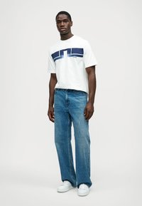 Balta medvilninė marškinė su tamsiai mėlynu grafiniu dizainu per krūtinę, derinama su džinsais ir baltais sportbačiais. Modelis stovi prieš neutralią foną.