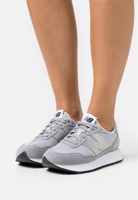 Zapatillas grises que combinan materiales de malla y ante, con una suela de goma negra, detalles en blanco y un diseño con costuras.