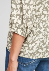 Poignet de manche d'un chemisier à fleurs vert et blanc avec un petit bouton, porté avec un jean en denim bleu.