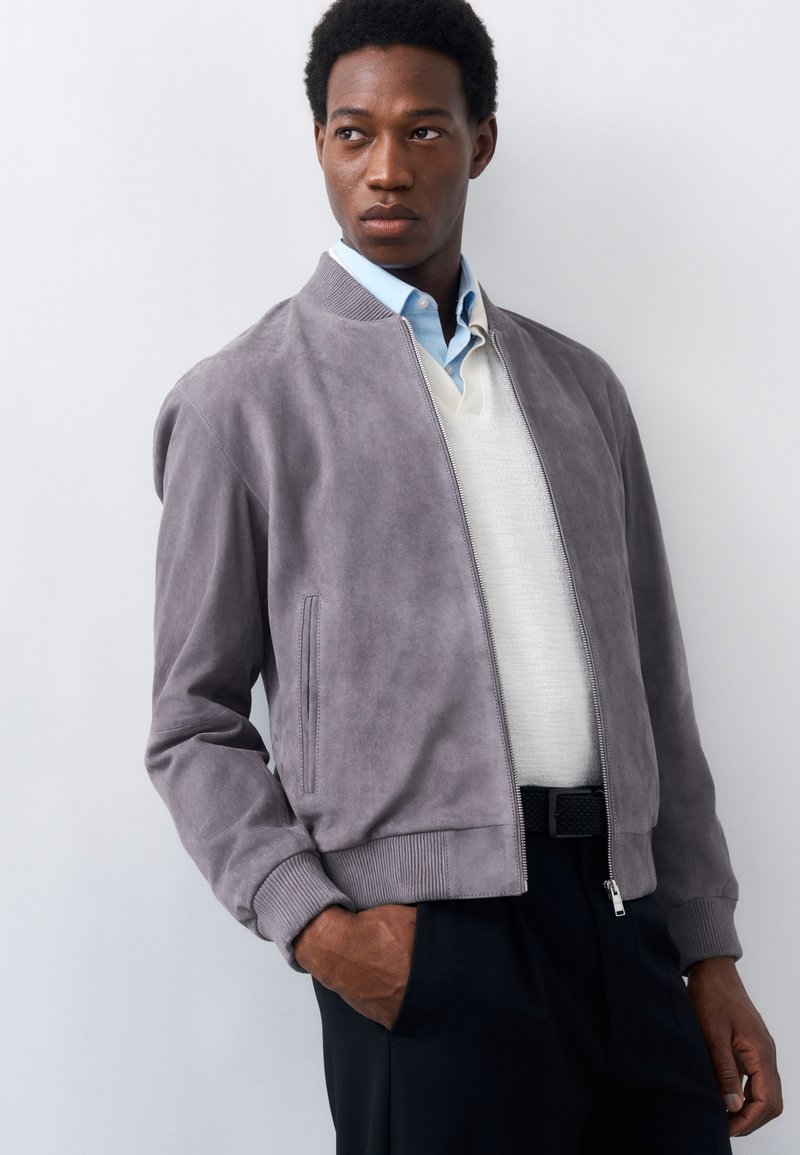 Homme portant une veste bomber en daim gris par-dessus un pull blanc et une chemise bleu clair à col, debout devant un fond clair uni.