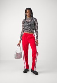 Calças de treino vermelhas com riscas laterais brancas, uma blusa de manga longa com padrão preto e branco, e uma bolsa metálica prateada. Sapatos negros.