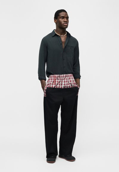 Άνδρας που φοράει σκούρο πουκάμισο με κουμπιά, κόκκινο και λευκό καρό στρώμα, μαύρο παντελόνι με φαρδιά μπατζάκια, μαύρα παπούτσια και κολιέ με κοχύλι, στέκεται με τα χέρια στις τσέπες.