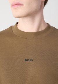 Gros plan d'une personne portant une chemise marron avec le mot BOSS imprimé en noir sur la poitrine.