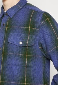 Kostkovaná flanelová košile v modré a zelené barvě s límečkem, zapínáním na knoflíky a dvěma náprsními kapsami se zapínáním na patentky. Texturovaná tkanina.