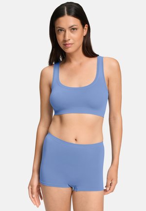 Donna con capelli lunghi e scuri che indossa un reggiseno sportivo blu abbinato e mutandine boyshort, in piedi davanti a uno sfondo semplice.