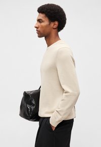 Jersey beige texturizado con escote redondo llevado por una persona, que sostiene un bolso negro acolchado, sobre un fondo claro.