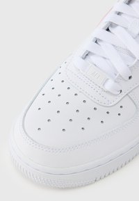 Biely kožený tenisk s hladkou textúrou, perforovanou špičkou, plochými bielymi šnúrkami a malou značkou „AF1“ na jazyku.