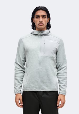 Hombre vestido con chaqueta con capucha gris clara con cremallera y bolsillo en el pecho, de pie mirando hacia adelante contra un fondo claro y liso.