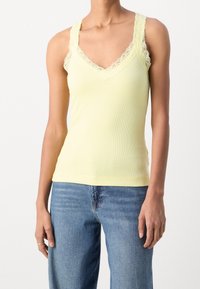 Kvinna iförd en ljusgul linne med spetskant och blå denimjeans, stående mot en enfärgad vit bakgrund.