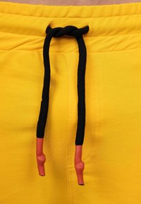 Gele joggingbroek van een katoenen mix met een zwarte trekkoord, voorzien van rode plastic aglets, en een gladde, zachte textuur.