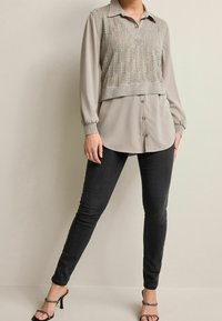 Gilet grigio a strati sopra una blusa grigio chiaro con bottoni, abbinato a jeans skinny neri e sandali con cinturini, caratterizzato da una texture liscia.