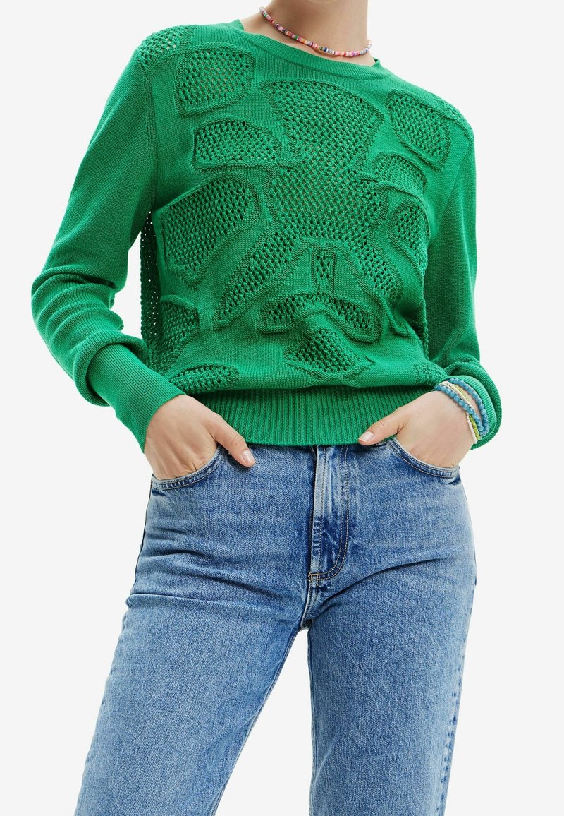 Personne portant un pull en maille vert avec des découpes géométriques, un jean bleu taille haute, un collier de perles et des bracelets, mains dans les poches.