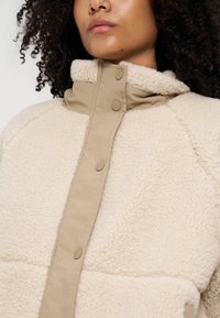 Personne portant une veste sherpa beige avec un col montant et des boutons-pression, montrant la partie supérieure du torse et la zone du cou.