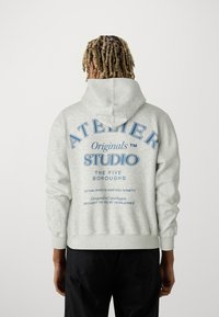 Pilkas džemperis su kauke, ant nugaros yra mėlynas užrašas: "ATELIER Originals STUDIO THE FIVE BOROUGHS." Minkšta tekstūra, standartinė iškirptė.