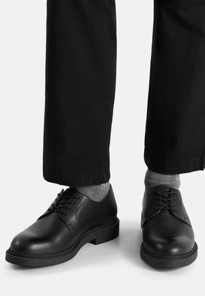 Schwarze Lederschuhe mit glatter Textur, rundem Zehenbereich und Schnürungsdetail, kombiniert mit grauen Socken und dunklen Hosen.