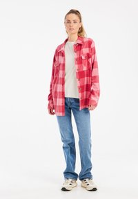 Camicia in flanella a quadri rossa e rosa con due tasche frontali, bottoni e maniche lunghe, indossata sopra una camicia bianca e abbinata a jeans blu.
