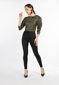 Oliven grüner, taillierter Lederoberteil mit Dreiviertelärmeln, kombiniert mit schwarzen, hochtaillierten Skinny-Hosen und schwarzen, spitzen Schuhen. Rote Quastenohrringe.