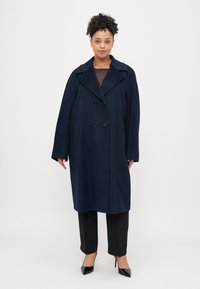 Cappotto in misto lana blu navy con design doppiopetto, ampi rever e tasche laterali. Indossato sopra una maglia nera e abbinato a pantaloni sartoriali.