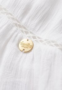 Witte stof met textuurdetails, voorzien van een ronde gouden metalen tag gegraveerd met "SuperDry" en "BRAND."