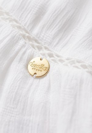 Tissu blanc avec des détails texturés, comportant une étiquette ronde en métal doré gravée "SuperDry" et "BRAND".