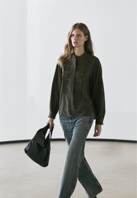 Frau geht drinnen und trägt eine dunkelgrüne Bluse mit langen Ärmeln, blaue Jeans und eine schwarze Patchwork-Einkaufstasche.