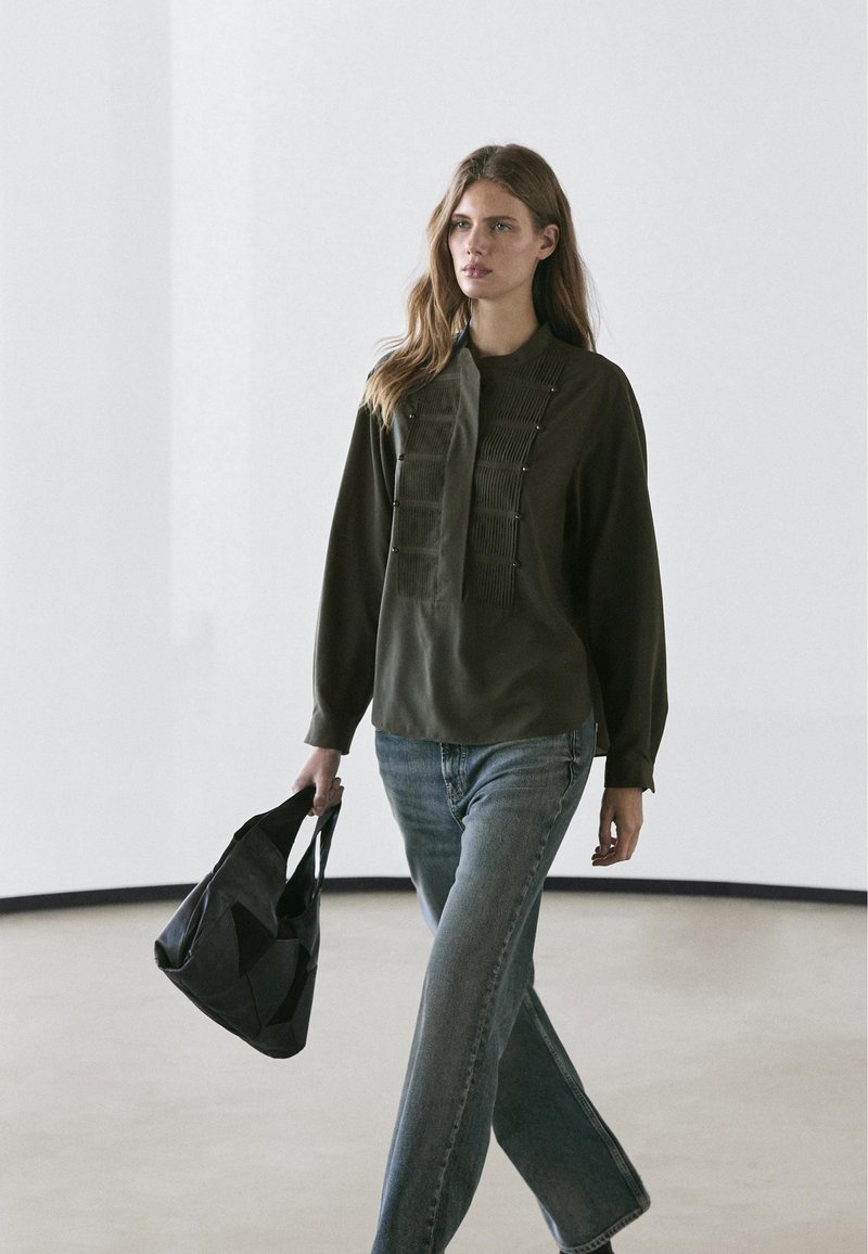 Frau geht drinnen und trägt eine dunkelgrüne Bluse mit langen Ärmeln, blaue Jeans und eine schwarze Patchwork-Einkaufstasche.