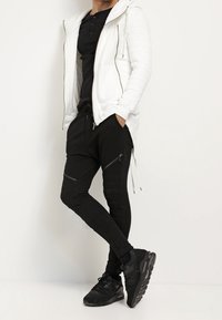 Bílá prošívaná mikina na zip, černé tričko s dlouhým rukávem, černé slim fit kalhoty se zipy a černé tenisky. Jednoduchý a moderní sportovní outfit.