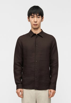 Dunkelbraunes Langarmhemd aus strukturiertem Stoff, mit durchgehendem Knopfverschluss, Kragen und geradem Saum, kombiniert mit hellbeigen Hosen.