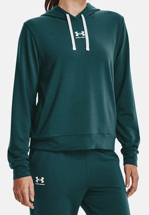 Person iført mørkegrøn Under Armour hættetrøje med hvide snører og matchende bukser, stående afslappet.