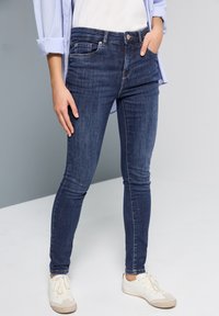 Jeans ajustados azul oscuro hechos de denim, con cinco bolsillos y un ajuste de altura media, combinados con zapatillas blancas sobre un fondo claro.