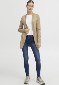 Beige, könnyű kardigán, nyitott elülső résszel és bordázott szegéllyel, fehér pólóval és sötétkék, skinny farmernadrággal párosítva. A megjelenést sportos cipők teszik teljessé.