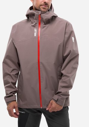 Man wearing taupe waterproof jacket with orange zipper and hood, featuring "Millet" logo on chest and sleeve.