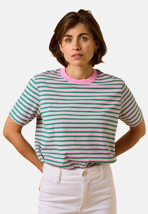 Eine Frau mit kurzen braunen Haaren trägt ein rosa-grün gestreiftes kurzärmeliges Shirt und weiße Hosen, steht mit den Händen hinter dem Rücken.